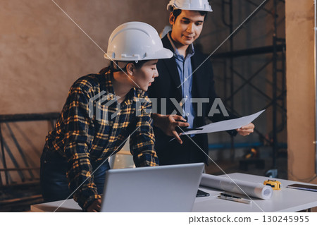 Construction manager and engineer dressed in orange work vests and hard helmets explore construction documentation on the building site near the steel frames Construction manager and engineer dressed in orange work vests and hard helmets explore construction documentation on the building site near the steel frames 130245955