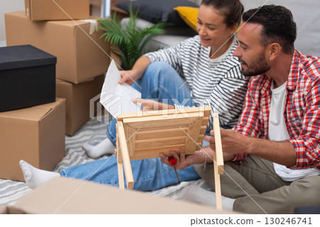 Home DIY: A young couple assembles a DIY shelf looking to paper instruction amidst moving carton boxes, adding a personal touch to their new home 130246741