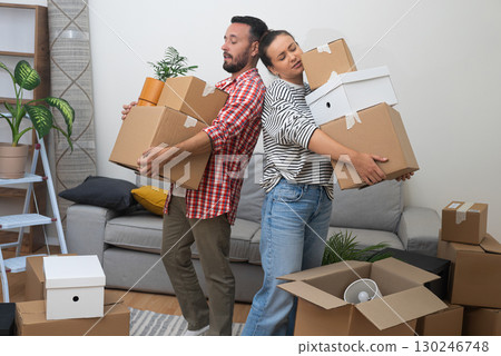 Moving Together: A young couple, proud homeowners, carries a heavy stack of cardboard relocation boxes into their new apartment's living room, embracing the move 130246748