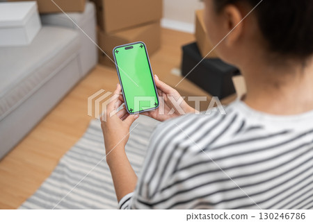 Mobile Relocation: Woman's hand with a smartphone, green screen in view, against a blurred backdrop of cardboard boxes, symbolizes a modern home move Mobile Relocation: Woman's hand with a smartphone, green screen in view, against a blurred backdrop of cardboard boxes, symbolizes a modern home move 130246786
