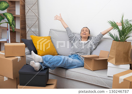 With a satisfied smile, a woman relaxes on her sofa amidst unpacked boxes, celebrating the successful transition to her new home during this joyful relocation 130246787