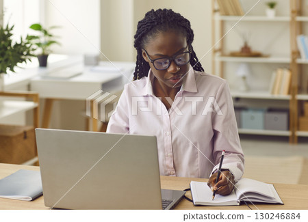 Serious african american business woman working on a laptop and making notes in office. Serious african american business woman working on a laptop and making notes in office. 130246884