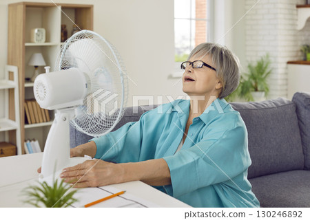 Senior woman, tired of extreme summer heat, enjoying fresh air by electric fan at home 130246892