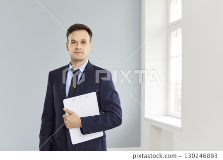 Man realtor agent standing in empty impartments with documents in hands and looking at camera Man realtor agent standing in empty impartments with documents in hands and looking at camera 130246893