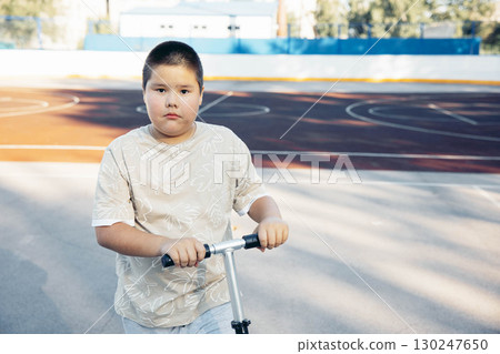 boy standing with scooter on outdoor sports court looking at camera with serious face, child in casual t shirt enjoying active lifestyle and childhood recreation, health, sport, childhood 130247650