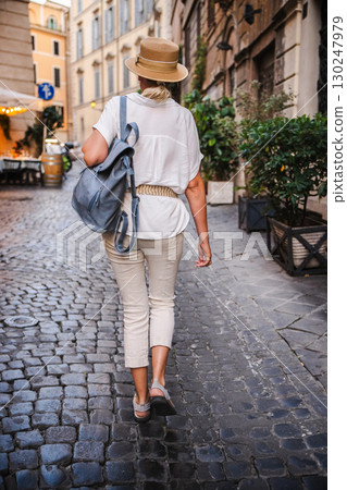 Middle-aged woman in a straw hat and light summer outfit with a backpack walking along a cobblestone street in Rome 130247979