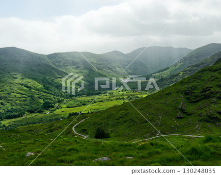 A landscape view shows green hills under an overcast sky. A winding road cuts through the lush valley floor in Gap of Dunloe, County Kerry, Ireland. 130248035