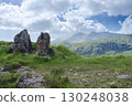 A picturesque landscape features grassy terrain with large rocks in the foreground, with mountainous hills and a cloudy sky visible in the distance, captured in Gap of Dunloe, Ireland. 130248038