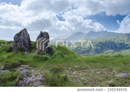 A picturesque landscape features grassy terrain with large rocks in the foreground, with mountainous hills and a cloudy sky visible in the distance, captured in Gap of Dunloe, Ireland. 130248038