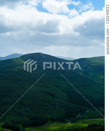 A tranquil landscape shows rolling green hills and valleys under a partly cloudy sky in the Gap of Dunloe, Ireland. The mountain and valley create a peaceful and scenic vista. 130248051