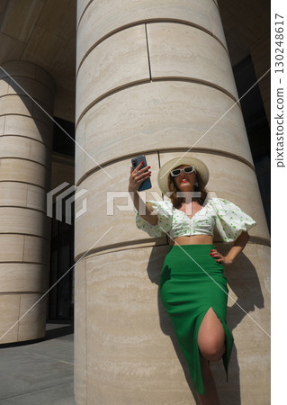 Beautiful tourist girl with hat holding smartphone and talking at the city square. 130248617