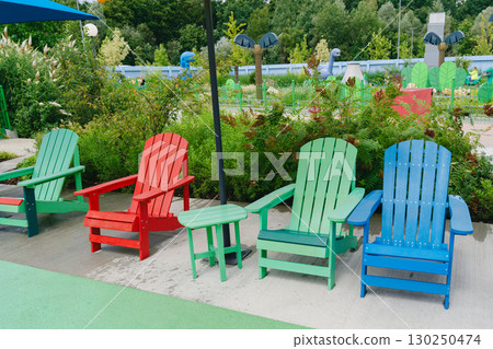 Vibrant red, green, and blue Adirondack chairs arranged in a park with lush greenery and a playground backdrop, creating a serene outdoor seating area for relaxation. 130250474