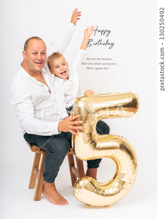 Joyful father and son sitting together, smiling and celebrating a 5th birthday with a golden balloon. Perfect for birthday themes, family love, parenting, and celebration concepts. Joyful father and son sitting together, smiling and celebrating a 5th birthday with a golden balloon. Perfect for birthday themes, family love, parenting, and celebration concepts. 130250492