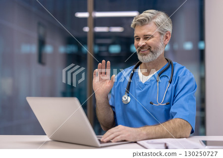 A professional doctor wearing scrubs participates in an online virtual meeting, smiling and waving. 130250727