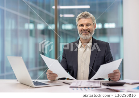 Professional man sitting at a desk, holding papers and smiling, in a modern workspace. 130250810
