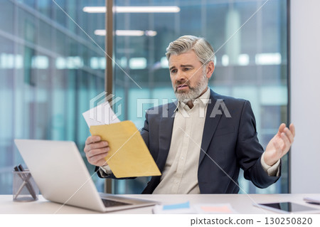 A mature businessman looks concerned while reviewing paperwork in a modern office setting. A mature businessman looks concerned while reviewing paperwork in a modern office setting. 130250820