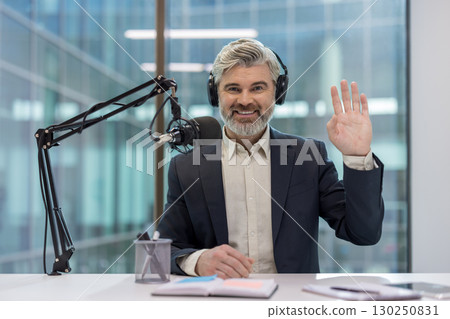 Confident male podcaster with headphones and microphone in a modern office environment, gesturing during a recording session. Professional broadcasting setting emphasizing communication 130250831