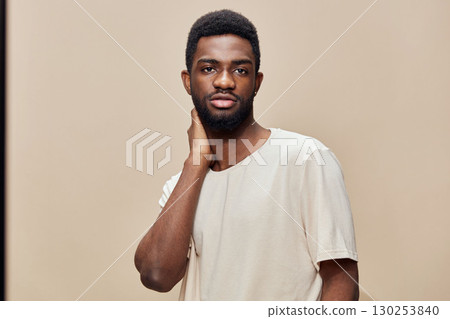 Confident young black man standing against a beige background, wearing a stylish neutral colored t shirt, expressing calm emotions and modern aesthetic 130253840