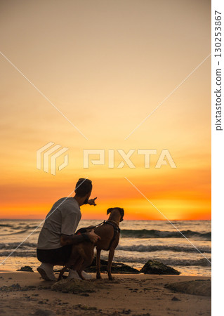 Man kneeling with his German Boxer dog on the beach at sunset, pointing towards the horizon while waves roll in, enjoying a quiet evening by the sea. Man kneeling with his German Boxer dog on the beach at sunset, pointing towards the horizon while waves roll in, enjoying a quiet evening by the sea. 130253867