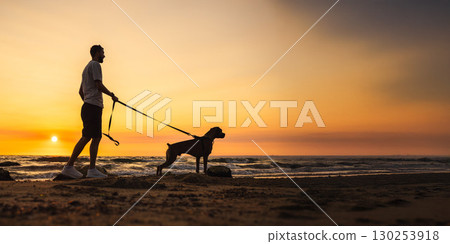 Man standing on the beach holding a German Boxer dog on a leash, both facing the ocean during a beautiful sunset, silhouette view with waves in the background. 130253918