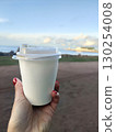 A woman's hand holds a paper cup of coffee against the backdrop of the Gulf of Finland. Close-up of a white cup with an empty space for a stamp. Russia, St. Petersburg, Peterhof National Park. 130254008