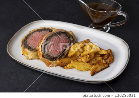 Beef Wellington with baked potato slice and demi-glace sauce. Traditional festive dish of beef tenderloin with mushrooms and Parma ham baked in puff pastry. Selective focus, close-up. Beef Wellington with baked potato slice and demi-glace sauce. Traditional festive dish of beef tenderloin with mushrooms and Parma ham baked in puff pastry. Selective focus, close-up. 130256031