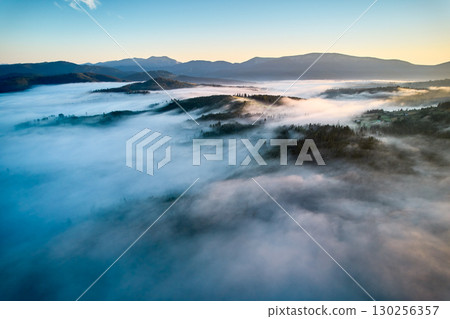 Breathtaking aerial view at dawn, showcasing vast sea of clouds enveloping mountain landscape. Subtle hues of rising sun paint sky in soft pastels, creating tranquil and ethereal scene. 130256357