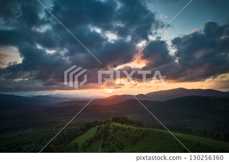 Aerial view breathtaking sunset over forest. Sun's rays break through dramatic clouds, casting golden glow across rolling hills and valleys. Distant mountains creating serene and picturesque scene. 130256360