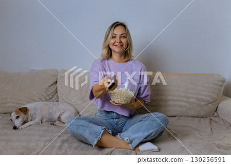 Happy young woman watching TV and smiling, holding popcorn bowl, sitting on sofa with cute dog. Cozy home evening, relaxing time with pet and favorite show. 130256591