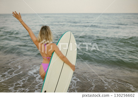 A woman carrying a surfboard walks toward the waves on a sunny beach, ready for a surfing session. This back view highlights her connection with the ocean and is perfect for themes of summer 130256602