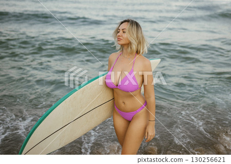 Attractive young woman in a purple bikini posing with a surfboard on the beach, enjoying summer by the sea, confident and relaxed, ocean waves in the background. Attractive young woman in a purple bikini posing with a surfboard on the beach, enjoying summer by the sea, confident and relaxed, ocean waves in the background. 130256621