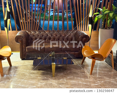 Leather sofa with wooden lounge chairs in hotel lobby. Interior decor, comfort and modern hospitality setting with vintage atmosphere. Leather sofa with wooden lounge chairs in hotel lobby. Interior decor, comfort and modern hospitality setting with vintage atmosphere. 130256647