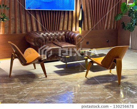 Elegant hotel lounge with leather sofa, wooden chairs and marble flooring. Retro inspired seating arrangement emphasizing hospitality, comfort and welcoming atmosphere. 130256678