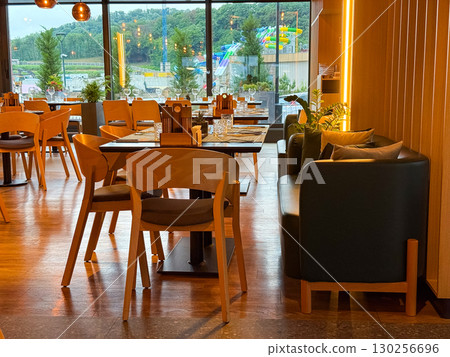 Restaurant interior with modern wooden chairs and decorative plants near wide glass windows. Dining space, hospitality, leisure and evening lifestyle atmosphere. Restaurant interior with modern wooden chairs and decorative plants near wide glass windows. Dining space, hospitality, leisure and evening lifestyle atmosphere. 130256696