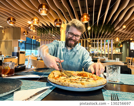 Man enjoying pizza while cutting slices with scissors in restaurant. Dining, leisure, lifestyle and modern food culture experience in casual interior with joyful atmosphere. Man enjoying pizza while cutting slices with scissors in restaurant. Dining, leisure, lifestyle and modern food culture experience in casual interior with joyful atmosphere. 130256697