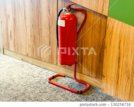 Red fire extinguisher standing on metal holder against wooden wall inside hotel corridor. Safety, emergency, and protection symbolizing equipment, hazard prevention, and workplace security. 130256716