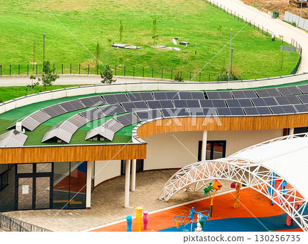 Building with solar panels installed on curved roof near green landscape and playground. Renewable energy, technology, and sustainability symbolizing innovation, ecology, and environmental balance. Building with solar panels installed on curved roof near green landscape and playground. Renewable energy, technology, and sustainability symbolizing innovation, ecology, and environmental balance. 130256735