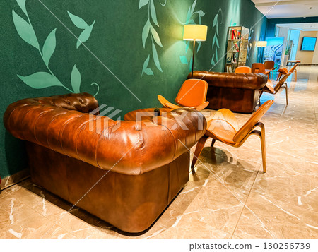 Leather sofas and wooden chairs arranged in modern lounge interior with soft lighting. Hospitality, comfort, and interior design symbolizing relaxation, lifestyle, and contemporary atmosphere. Leather sofas and wooden chairs arranged in modern lounge interior with soft lighting. Hospitality, comfort, and interior design symbolizing relaxation, lifestyle, and contemporary atmosphere. 130256739