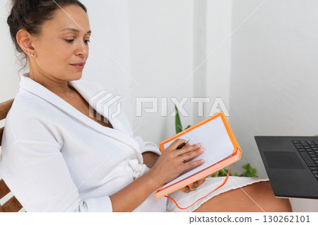Woman in White Robe Writing Notes at Laptop in Bright Minimalistic Space 130262101