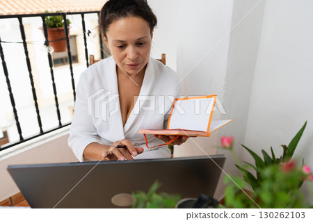 Woman Working Remotely with a Laptop and Notebook on a Sunny Balcony 130262103