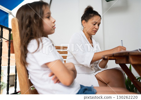 Mother Focusing on Work While Child Sits Nearby on a Sunny Balcony 130262220