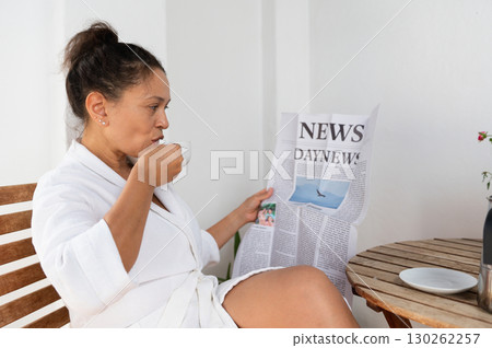 Woman in White Bathrobe Enjoying Coffee and Reading Newspaper Outdoors Woman in White Bathrobe Enjoying Coffee and Reading Newspaper Outdoors 130262257