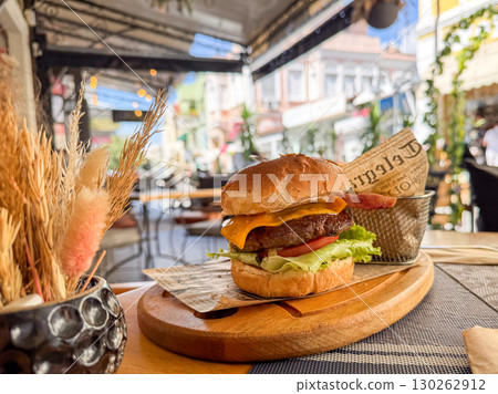 A juicy cheeseburger with bacon, lettuce, and tomato served on a wooden plate in an outdoor cafe 130262912
