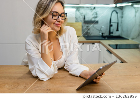 Relaxed woman in glasses using digital tablet in modern kitchen. Morning routine, watching news or browsing content, casual home atmosphere, elegant lifestyle. 130262959