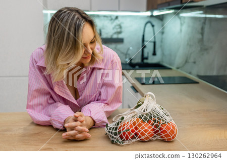 Smiling woman in pink shirt leaning on kitchen counter next to mesh bag with fresh vegetables. Healthy lifestyle and eco-friendly food shopping at home. 130262964