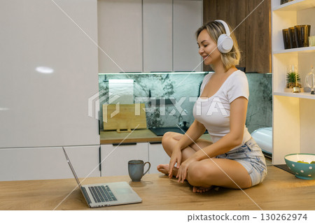 Smiling young woman in headphones sitting on kitchen counter with laptop. Modern cozy interior, casual home scene, digital lifestyle, evening relaxation in contemporary kitchen. Smiling young woman in headphones sitting on kitchen counter with laptop. Modern cozy interior, casual home scene, digital lifestyle, evening relaxation in contemporary kitchen. 130262974