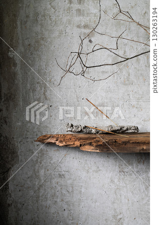Burning incense stick on a rustic holder with ash, placed against a textured concrete wall with dry branches and old wood. Minimalist still life in wabi-sabi style. 130263194