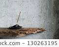 Burning incense stick on a rustic holder with ash, placed against a textured concrete wall with dry branches and old wood. Minimalist still life in wabi-sabi style. 130263195