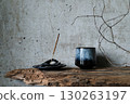 Burning incense stick in a hand-shaped holder with a ceramic cup on rustic wood against a concrete wall. Minimalist wabi-sabi still life. 130263197