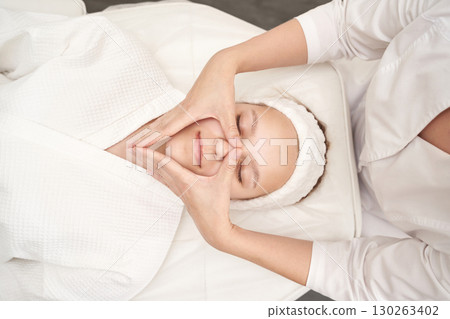 Young caucasian female receiving facial massage in spa wearing white robe 130263402